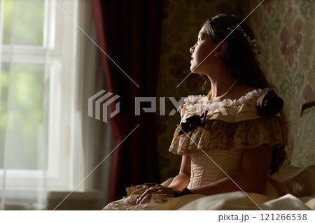 Side view of girl in classic gown gazing thoughtfully out window in softly illuminated vintage room, copy space 121266538