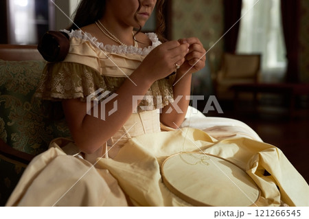 Cropped shot of young lady wearing classic dress with ruffles and frills while starting new embroidery and threading needle, copy space Cropped shot of young lady wearing classic dress with ruffles and frills while starting new embroidery and threading needle, copy space 121266545
