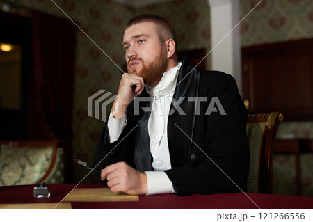 Portrait of young bearded gentleman wearing tailcoat looking away pensively while daydreaming at table in classic interior 121266556