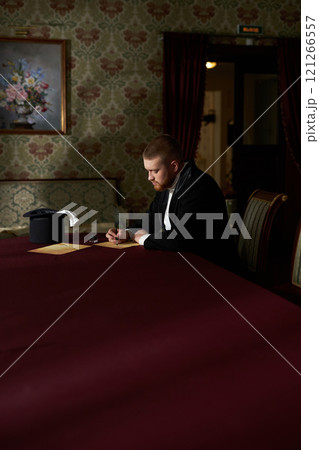 Vertical wide angle portrait of young man wearing tuxedo writing letter while sitting at table alone in classic interior with natural lighting 121266557