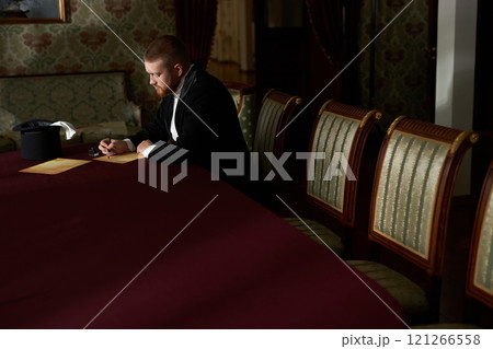 Wide angle image of young gentleman writing letter with ink while sitting at red velvet table in classic interior, copy space 121266558