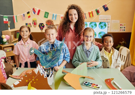 Front view portrait of happy teacher posing with multiethnic group of children holding handmade dinosaur crafts in colorful art classroom copy space Front view portrait of happy teacher posing with multiethnic group of children holding handmade dinosaur crafts in colorful art classroom copy space 121266579