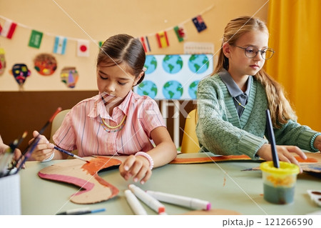 Portrait of two young girls painting dinosaur figures made of cardboard while enjoying creative activity in colorful classroom copy space 121266590