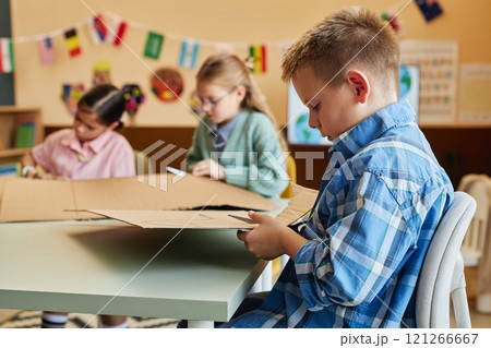 Side view portrait of blonde young boy cutting cardboard while enjoying art and craft class for children copy space Side view portrait of blonde young boy cutting cardboard while enjoying art and craft class for children copy space 121266667
