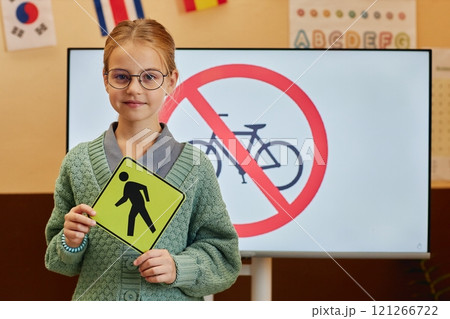 Waist up portrait of young girl holding pedestrian crossing sign and looking at camera during road safety lesson for children copy space 121266722