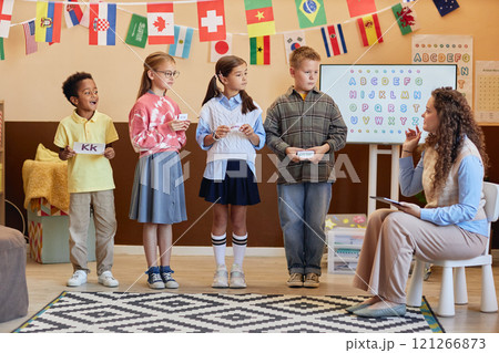 Full length view of diverse group of young children holding alphabet cards standing in colorful primary school classroom and learning English with female teacher Full length view of diverse group of young children holding alphabet cards standing in colorful primary school classroom and learning English with female teacher 121266873