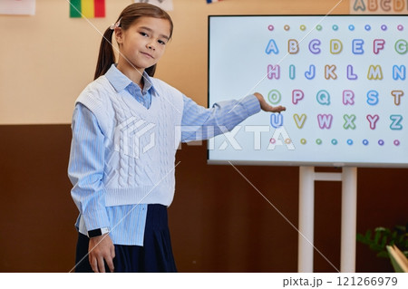 Waist up of portrait of cute young girl pointing at digital screen with colorful alphabet letters while learning English in primary school 121266979