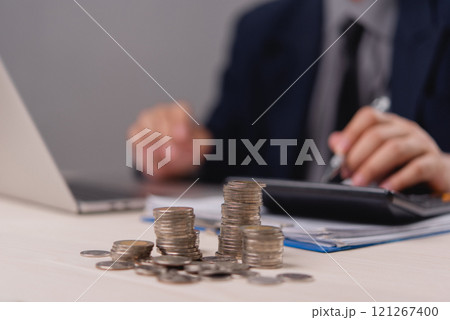 Stacks of coins with businessman calculating finances on desk in the background representing investment and saving Stacks of coins with businessman calculating finances on desk in the background representing investment and saving 121267400