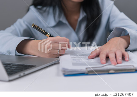 Close-up of woman writing on documents with pen beside a laptop 121267407
