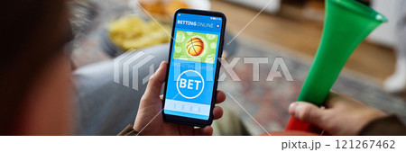 Header image of man holding smartphone with online sports bets app on screen while watching basketball game at home, copy space Header image of man holding smartphone with online sports bets app on screen while watching basketball game at home, copy space 121267462