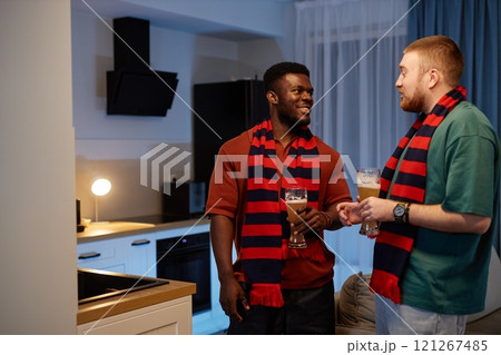 Side view portrait of two sports fans wearing matching team scarves talking and holding drinks discussing match in relaxed home setting, copy space 121267485
