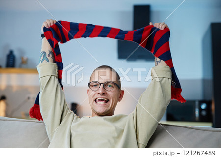Waist up portrait of excited sports fan cheering while holding striped scarf in team colors and watching match on TV at home, copy space Waist up portrait of excited sports fan cheering while holding striped scarf in team colors and watching match on TV at home, copy space 121267489