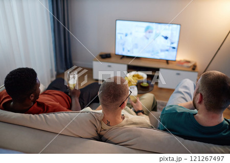 High angle back view at three friends sitting on couch with beer glasses and watching TV enjoying evening at home together, copy space High angle back view at three friends sitting on couch with beer glasses and watching TV enjoying evening at home together, copy space 121267497