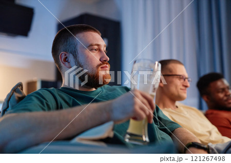 Side view portrait of bearded young man holding beer glass while watching TV with friends at home and holding beer glass, copy space Side view portrait of bearded young man holding beer glass while watching TV with friends at home and holding beer glass, copy space 121267498