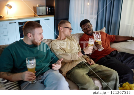 Portrait of three male friends clinking beer glasses while sitting on couch together and watching TV at home Portrait of three male friends clinking beer glasses while sitting on couch together and watching TV at home 121267513