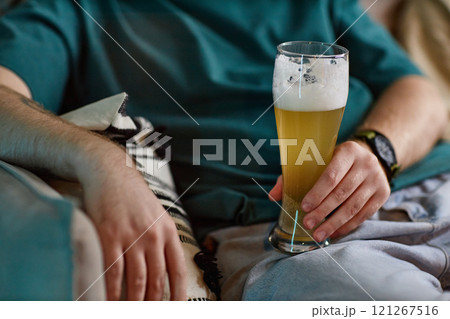 Close up of unrecognizable man holding frothy beer glass while watching TV at home lounging on couch, copy space Close up of unrecognizable man holding frothy beer glass while watching TV at home lounging on couch, copy space 121267516