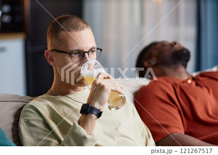 Portrait of adult man wearing glasses and drinking beer from tall glass while watching TV on couch with friends, copy space Portrait of adult man wearing glasses and drinking beer from tall glass while watching TV on couch with friends, copy space 121267517