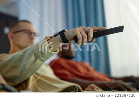 Side view close up of male hand pointing TV remote and switching channels sitting on comfortable couch with friends, copy space Side view close up of male hand pointing TV remote and switching channels sitting on comfortable couch with friends, copy space 121267520