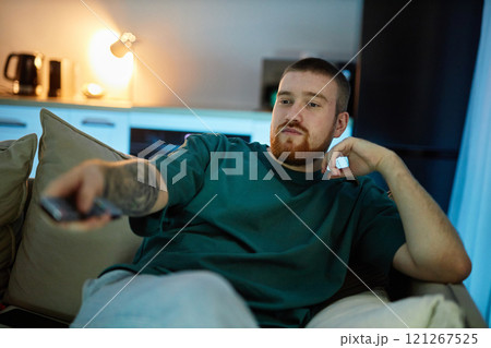 Front view portrait of relaxed bearded young man lounging on couch and holding TV remote in dimly lit living room, copy space 121267525