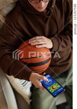 Vertical above view of young man placing bet on sports game online and holding basketball ball while watching game from home Vertical above view of young man placing bet on sports game online and holding basketball ball while watching game from home 121267586