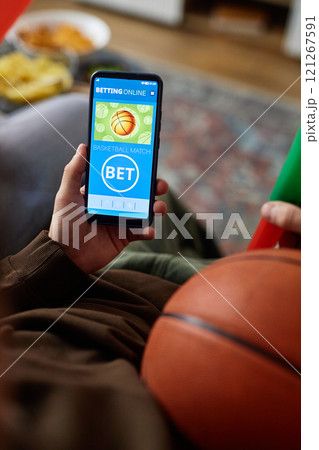 Vertical close up of unrecognizable man holding basketball and smartphone with sports betting app on screen over blurred background, copy space 121267591