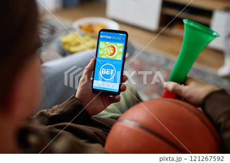 Close up unrecognizable man holding smartphone with online sports bets app on screen while watching basketball game at home, copy space 121267592