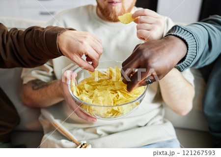 Front view closeup of diverse male hands grabbing potato chips from clear glass bowl during intense game moment on TV, copy space 121267593