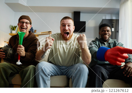 Front view portrait of three joyful men sitting together on couch yelling with excitement while watching sports game on TV and holding sports fan accessories Front view portrait of three joyful men sitting together on couch yelling with excitement while watching sports game on TV and holding sports fan accessories 121267612