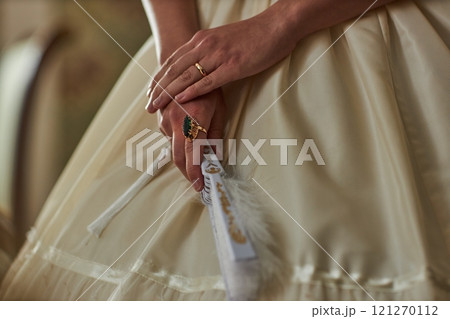 Close up detail shot of ladys hands with emerald ring holding ornate lace fan over soft cream dress, copy space 121270112