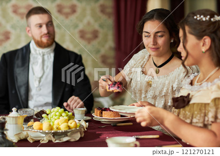 Side view portrait of young gentleman in velvet tailcoat looking at elegant ladies at dining table in classic interior lit by warm lighting Side view portrait of young gentleman in velvet tailcoat looking at elegant ladies at dining table in classic interior lit by warm lighting 121270121