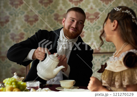 Portrait of classic young gentleman in black tailcoat smiling while pouring tea for young lady sitting at table in vintage setting 121270129