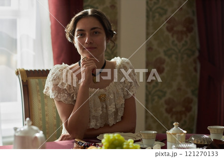 Front view portrait of classic young lady in lace dress sitting gracefully at table and looking at camera resting chin on hand with soft natural lighting, copy space 121270133