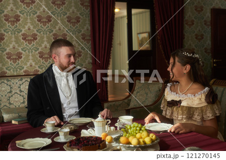 Romantic scene of young lady and gentleman dressed in vintage attire sitting at tea table smiling at each other in classic interior 121270145