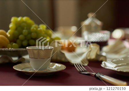 Close up of single tea cup and saucer with blurred fruit and cutlery in background on crimson vintage table, copy space 121270150