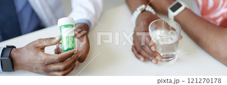 Detail image of African American doctor holding green pill bottle with patient holding glass of water and taking medication in clinic, copy space Detail image of African American doctor holding green pill bottle with patient holding glass of water and taking medication in clinic, copy space 121270178