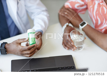 Close up of African American doctor holding green pill bottle with patient holding glass of water and taking medication in clinic, copy space 121270518
