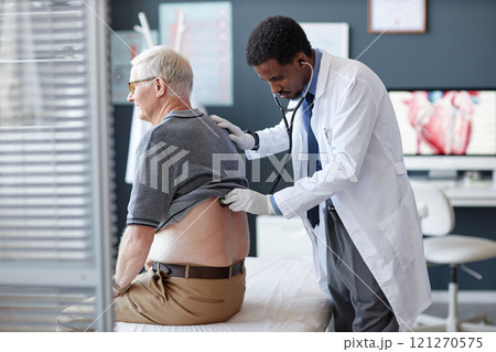 Side view of African American doctor using stethoscope and listening to heart and breathing while examining senior patient during checkup in clinic, copy space 121270575