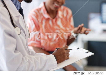 Side view close up of unrecognizable African American doctor writing notes on clipboard with woman explaining symptoms in blurred background, copy space 121270583