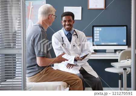 Waist up portrait of smiling African American doctor wearing lab coat and holding tablet while talking to senior patient during consultation in clinic, copy space Waist up portrait of smiling African American doctor wearing lab coat and holding tablet while talking to senior patient during consultation in clinic, copy space 121270591