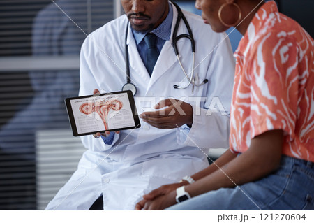 Cropped shot of African American male OBGYN wearing lab coat and holding tablet displaying female reproductive system diagram while talking to young woman Cropped shot of African American male OBGYN wearing lab coat and holding tablet displaying female reproductive system diagram while talking to young woman 121270604