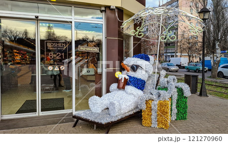 New Year's snowman on a sun lounger under an umbrella. A decorative snowman with a glowing umbrella was placed in front of the entrance to the store. Pre-holiday interior decorations. New Year's snowman on a sun lounger under an umbrella. A decorative snowman with a glowing umbrella was placed in front of the entrance to the store. Pre-holiday interior decorations. 121270960