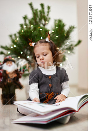 Cute little child girl reads a book near the Christmas tree at home Cute little child girl reads a book near the Christmas tree at home 121271047