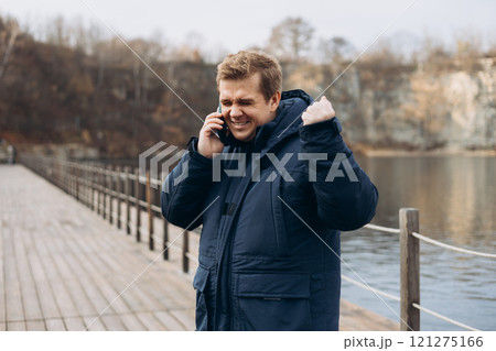 Male standing on the lake wooden pier. Happy man talk phone, walking near river. Phone Communication. Happy won the game, lottery, shows yes with his hand. Male standing on the lake wooden pier. Happy man talk phone, walking near river. Phone Communication. Happy won the game, lottery, shows yes with his hand. 121275166