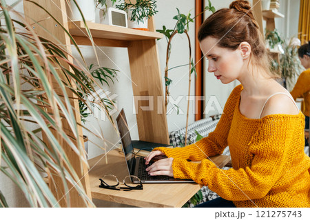 happy young woman is sitting at desk in home office, looking at laptop screen studying 121275743