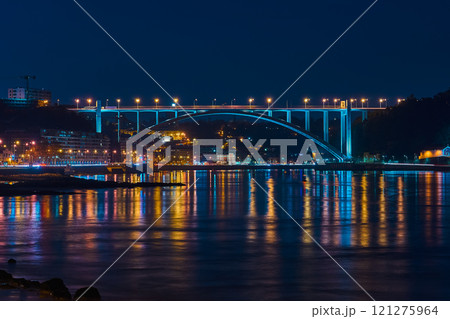 Lit bridge at night casting colorful reflections on calm river, surrounded by glowing city lights and dark urban skyline 121275964