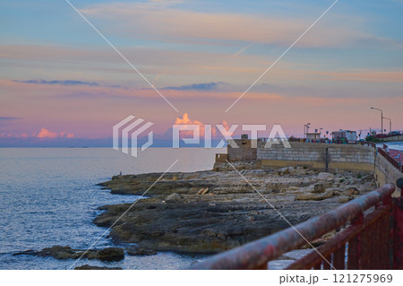 Coastal promenade featuring rocky shoreline under pastel-colored sunset sky, with calm sea waters stretching into horizon Coastal promenade featuring rocky shoreline under pastel-colored sunset sky, with calm sea waters stretching into horizon 121275969