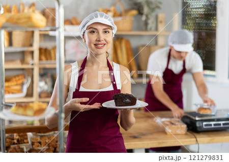Cheerful female baker offering slice of chocolate cake Cheerful female baker offering slice of chocolate cake 121279831
