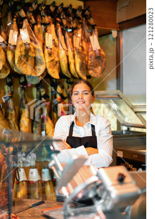 Portrait of positive female butcher on background of rack with hanging various jamon Portrait of positive female butcher on background of rack with hanging various jamon 121280023