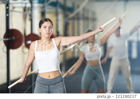 Slender girl, together with other athletes, does a warm-up using a long stick in gym Slender girl, together with other athletes, does a warm-up using a long stick in gym 121280217