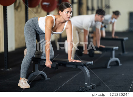 Athletic young woman pumping her biceps with a dumbbell while leaning on bench 121280244